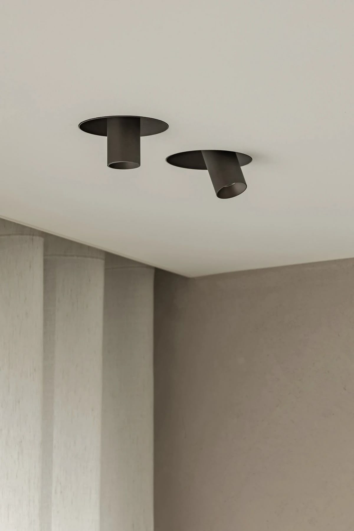 Two recessed bronze ceiling spotlights above a plastered wall and linen curtains